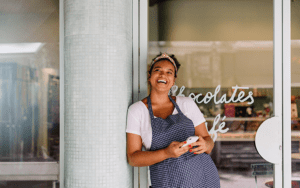 a woman in an apron holds a phone in her hand. She is standing in front of her cafe