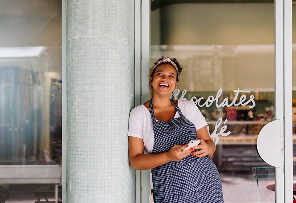 a woman in an apron holds a phone in her hand. She is standing in front of her cafe