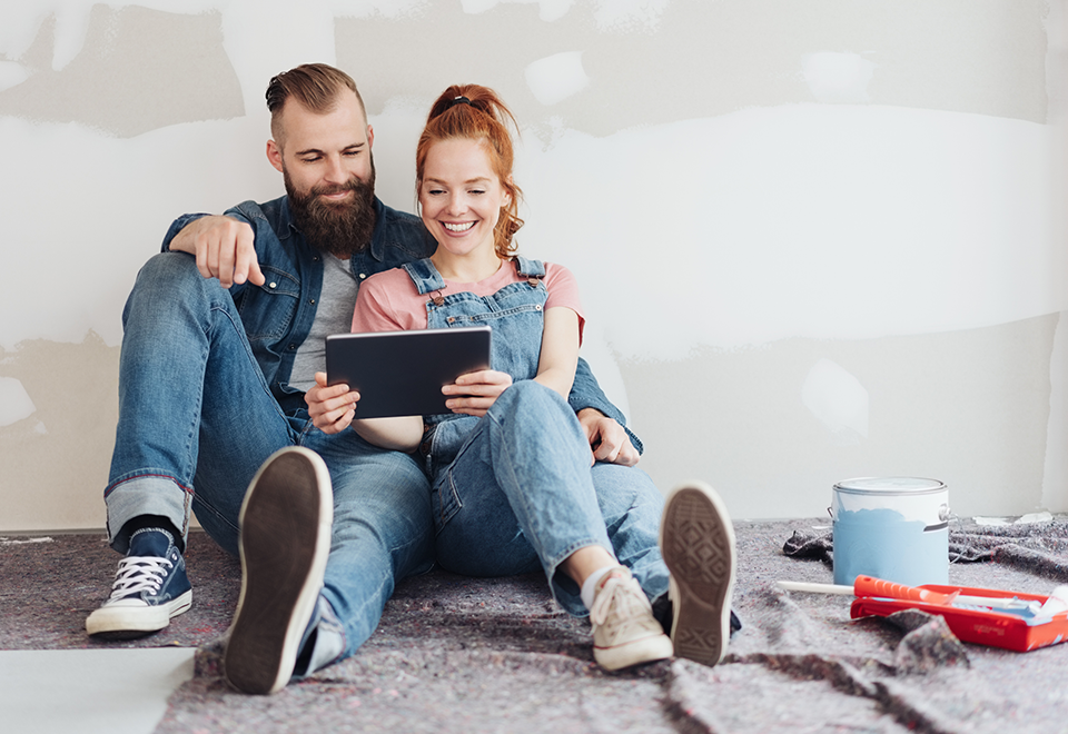 A couple is seated in front of their unfinished wall, holding a tablet. There are cans of paint and supplies next to them