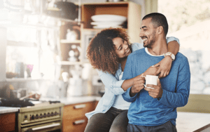 A couple is in the kitchen. The man is holding a cup of coffee and his wife has her arms wrapped around his neck, looking at his face lovingly