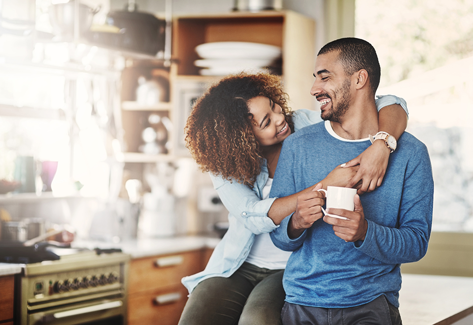 A couple is in the kitchen. The man is holding a cup of coffee and his wife has her arms wrapped around his neck, looking at his face lovingly