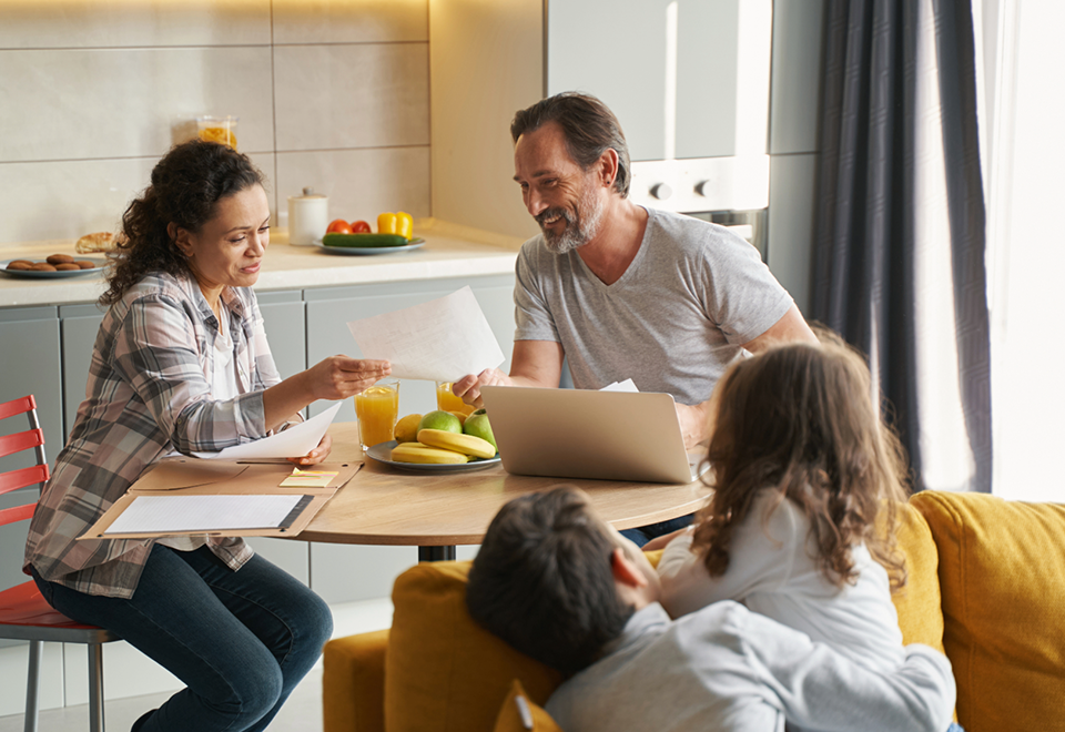 A husband and wife sit at the kitchen table looking through paper work while their children play nearby on the couch