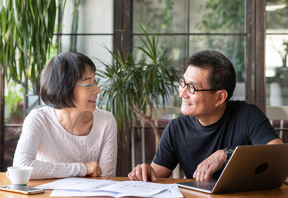 A man and a woman sit at a table with paperwork and a computer in front of them