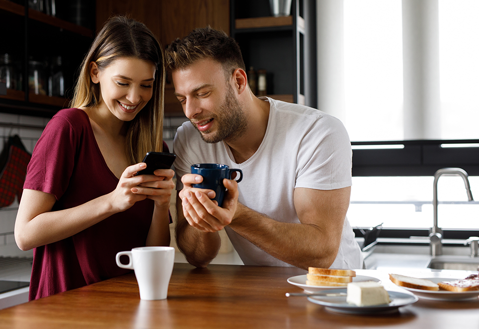 A man and woman sit at the breakfast table. He is holding a cup of coffee and she's holding her phone and they are both looking at her phone