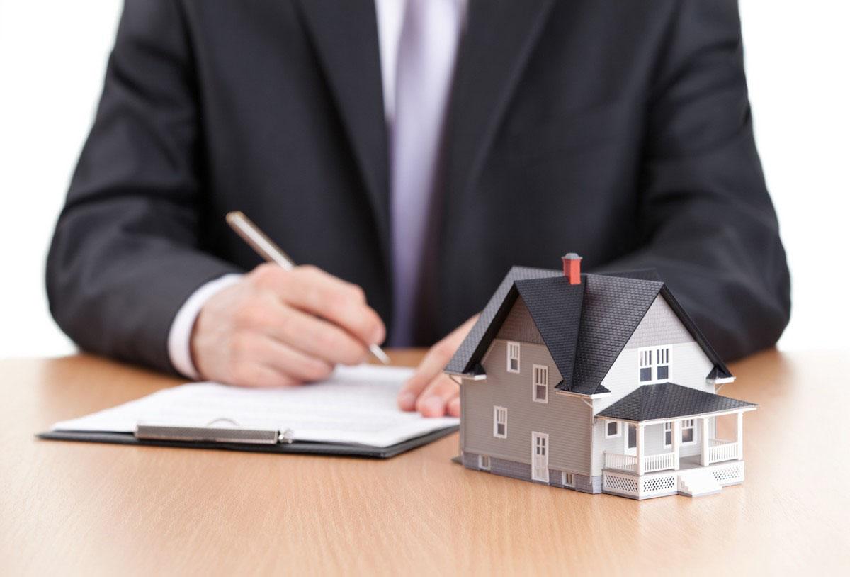 Photo of a man in a suit signing paper work. There is a model of a home in front of it