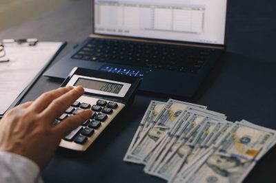 Person using a calculator beside a laptop displaying a spreadsheet with stacks of 100-dollar bills on the desk, counting money.