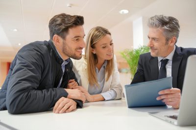 Three coworkers (two men, one woman) lean over a white table, examining a laptop and a blue document in a bright office.