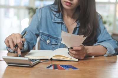 Person in a denim jacket writing with a pen while reviewing papers at a desk, with a calculator and cards nearby on the table.